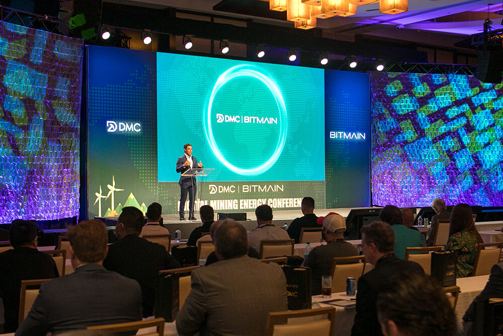 A man is speaking on stage in front of the Bitmain logo with an audience arranged in classroom style seating looks on