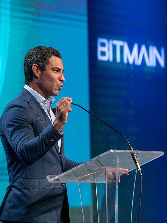 A close up shot of a man speaking at a podium with a blurry Bitmain logo in the background