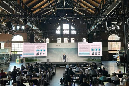 A man standing on a stage flanked by two large LED screens in an industrial space. Audience is sat a tables in classroom style set-up in the foreground