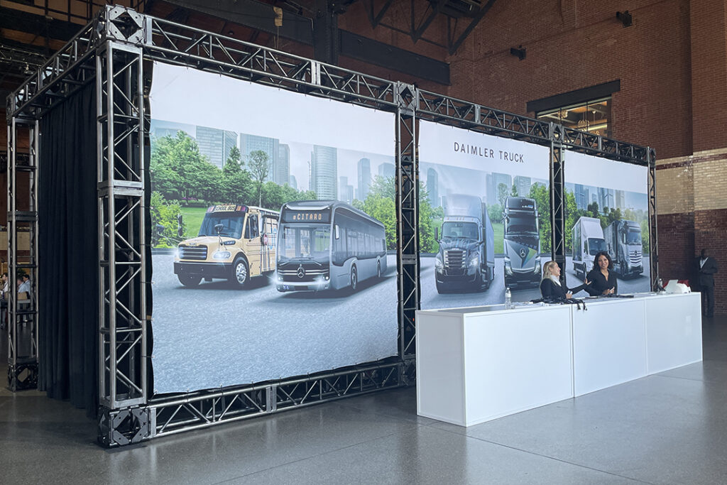 Two women talk at white counter in front of a Daimler Truck graphic wall supported by a truss structure.