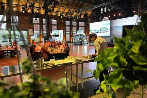 A chef is plating food in the foreground. Three large tables of guests and two semi-truck cabs are in the background.